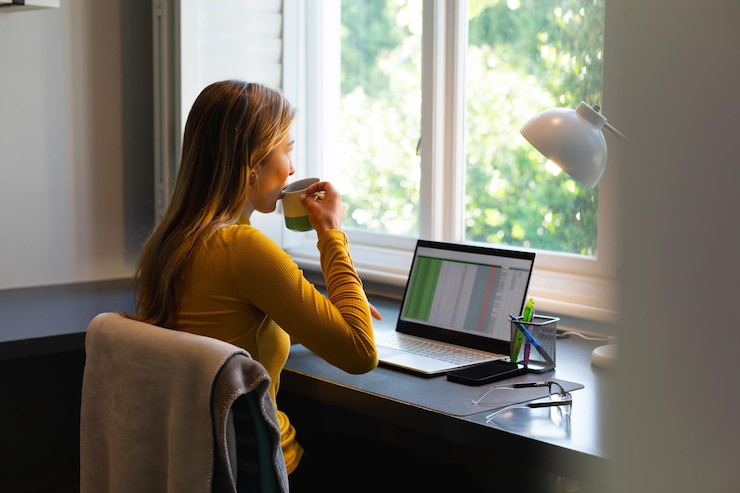 Foto weiße frau sitzt an einem schreibtisch, arbeitet von zu hause mit laptop und trinkt kaffee in einem sonnigen raum
