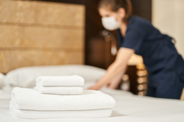 Foto weiße frische handtücher auf dem bett mit einem dienstmädchen in einer maske im hintergrund. hotelservicekonzept