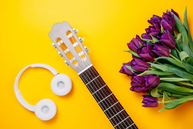 Weiße Gitarre und weiße kabellose Kopfhörer auf dem orangefarbenen Hintergrund, Frühlingsblumen, Bündel lila Tulpen, weiße Gitarre und Blumen, Frühlingsmusikplakat.