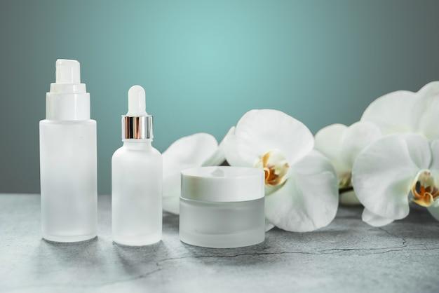 Weiße Glasserumflasche und Cremedose im Badezimmer mit Orchideenblüten im Hintergrund, markenlose Kosmetikprodukte, Spa-Kosmetikprodukt-Branding-Mockup