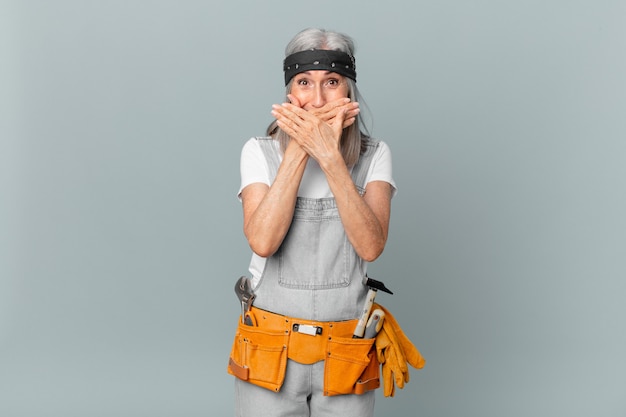 Weiße Haarfrau mittleren Alters, die den Mund mit den Händen mit einer schockierten und tragenden Arbeitskleidung und Werkzeugen bedeckt. Housekeeping-Konzept
