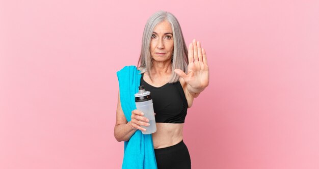 Weiße Haarfrau mittleren Alters, die ernst aussieht und offene Handfläche zeigt, die eine Stoppgeste mit einem Handtuch und einer Wasserflasche macht. Fitnesskonzept