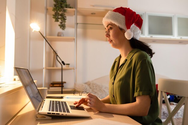 Weiße junge Frau mit Weihnachtshut arbeitet am Laptop Seitenansicht Konzept der Fernarbeit und