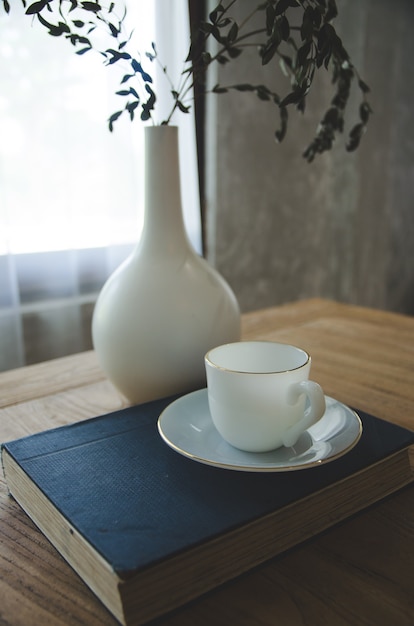 Weiße Kaffeetasse auf blauem Buch mit weißem Vase auf Tabelle