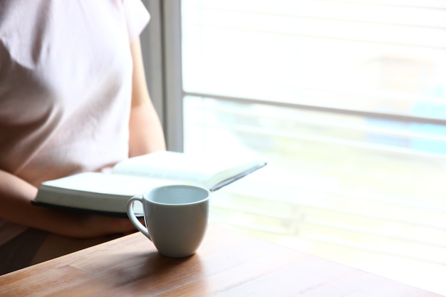 weiße Kaffeetasse auf dem Schreibtisch mit einer Frau, die ein Buch liest