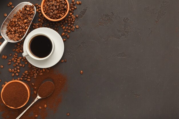 Weiße Kaffeetasse auf Untertasse und verstreute geröstete Bohnen auf grauem strukturiertem Hintergrund, Draufsicht. Minimalistisches Design für Coffeeshop-Werbung, Kopierraum