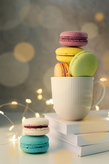 Weiße Kaffeetasse gefüllt mit Makronen auf dem Tisch, Lichterketten und Bokeh-Hintergrund