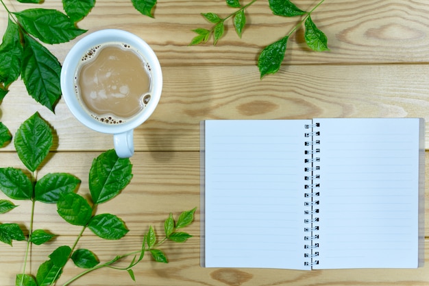 Weiße Kaffeetasse, grüne Blätter der Niederlassungen und Anmerkungsbuch auf einem Holztisch.