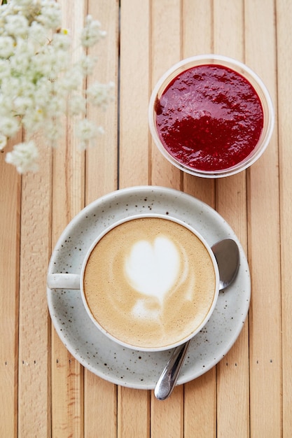 Weiße Keramiktasse Cappuccino mit Herz Himbeermarmelade Dessert ein Snack leichtes Frühstück auf der Caféterrasse mit verschwommenem Vordergrund aus Kamillenblüten Flaches Essen Vertikales Foto