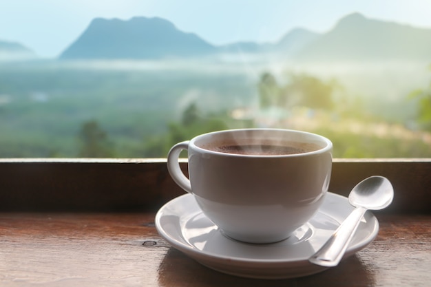 Weiße keramische Kaffeetasse auf Holztisch am Morgen mit Sonnenlicht über verschwommener Gebirgslandschaft