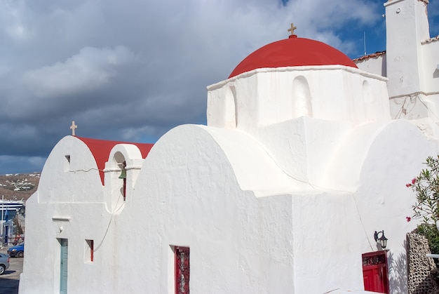 Weiße Kirche in Mykonos, Griechenland. Kirche mit roter Kuppel am bewölkten blauen Himmel. Kapellengebäudearchitektur auf sonnigem im Freien. Religion und Kultkonzept. Sommerurlaub auf der Mittelmeerinsel, Fernweh.