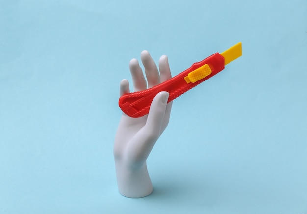 Weiße Schaufensterpuppe Hand hält Spielzeug-Papiermesser auf blauem Hintergrund. Renovierungskonzept, Industrie. Minimalismus