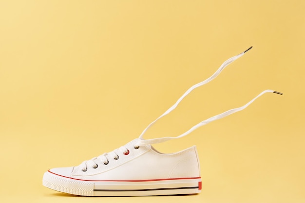 Weiße Sportturnschuhe für Damen mit fliegenden Schnürsenkeln vor gelbem Hintergrund Lässige trendige Schuhe Nahaufnahme Kreatives minimales Design mit Kopierraum