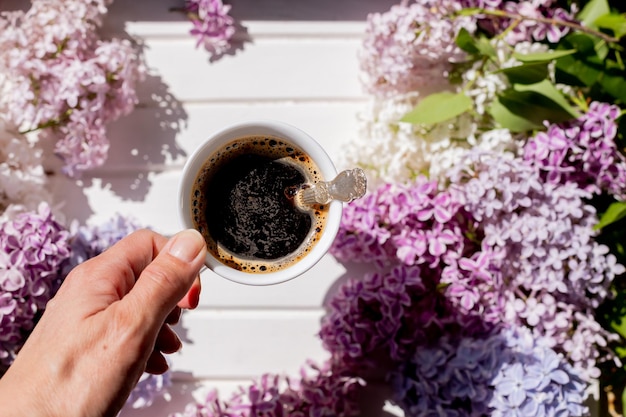 Weiße Tasse frischer Schwarzkaffee auf weißem Holztisch mit lila Flieder-Zweig-Morgenroutine-Kaffee