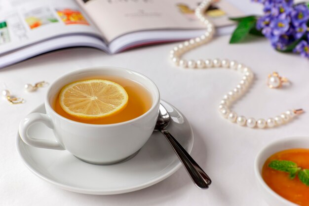 Weiße Tasse grüner Tee mit Zitrone, Honig und Minze auf einem weißen Bettlaken mit illustriertem Magazin, Perlenkette, Ohrringen und Ring.