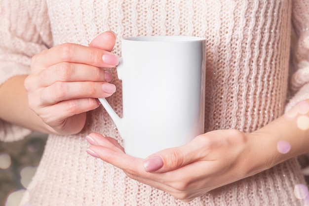 - Weiße Tasse in den Händen der Frau in einem rosa Winterpullover. Weihnachtskaffeetasse.