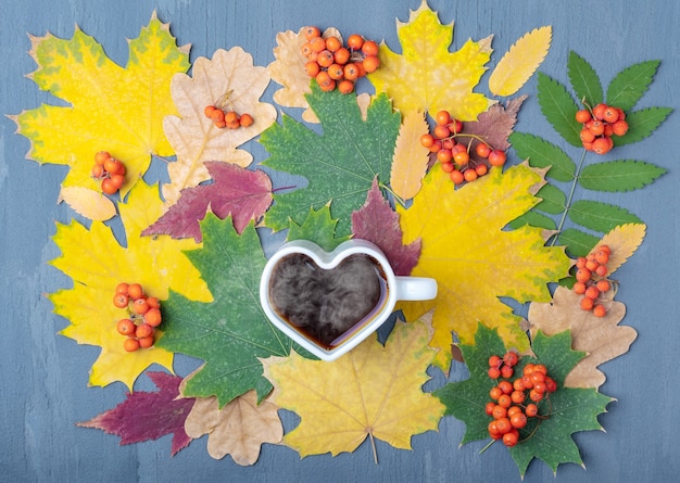 Weiße Tasse in Form eines Herzens mit dampfendem Kaffee oder Tee auf einem Hintergrund von Herbst gefallenen trockenen bunten Blättern und orange Vogelbeeren. Herbst-Blues-Konzept. Herbstdepression.