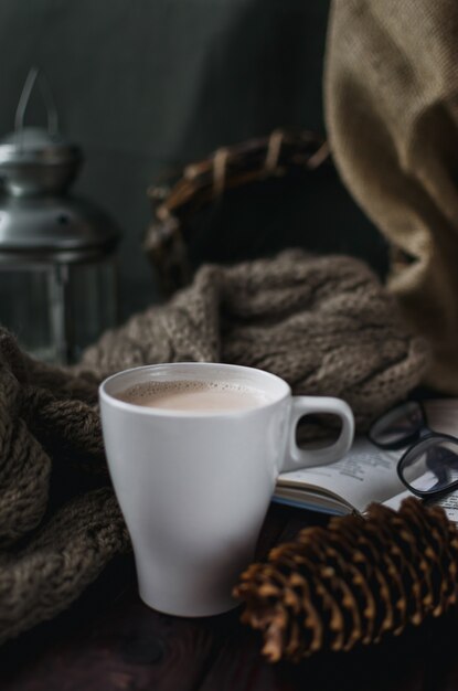 Weiße Tasse Kakao auf einem alten Holztisch mit einem unterhaltsamen Buch und einer Lesebrille.