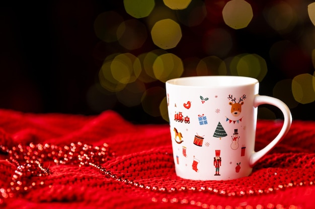 Weiße Tasse mit Neujahrsmuster auf einer roten Strickdecke auf dunklem Hintergrund an der Seite, Weihnachtsstillleben. Foto in hoher Qualität