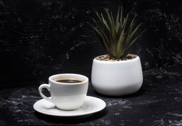 Weiße Tasse mit schwarzem aromatischem Kaffee auf einem schwarzen abstrakten Hintergrund mit einer Topfblume im Hintergrund.