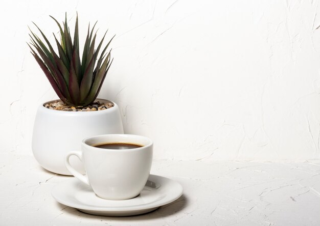 Weiße Tasse mit schwarzem aromatischem Kaffee auf einem weißen abstrakten Hintergrund mit einer Topfblume im Hintergrund mit einer Kopie des Raumes.