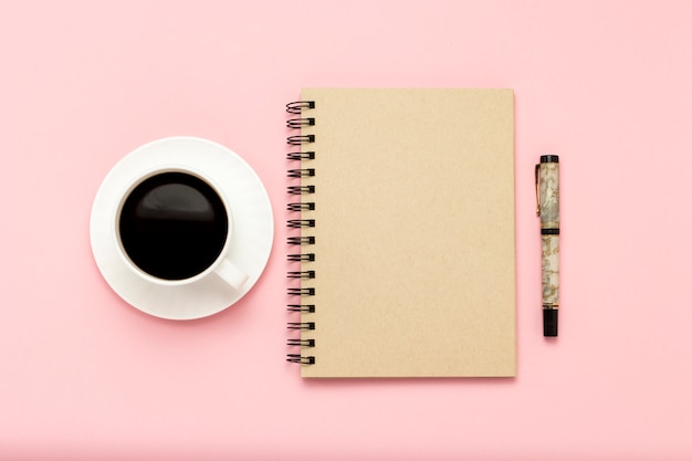 Weiße Tasse mit schwarzem Kaffee, Notizbuch, Stift auf einem rosa Hintergrund. Flache Lage, Draufsicht