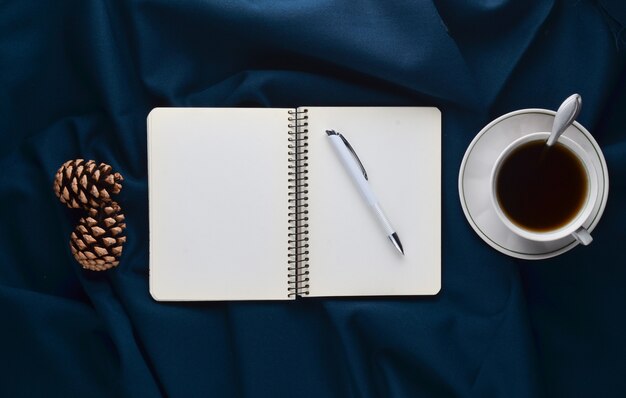 Weiße Tasse mit Tee, Notizbuch und Stift, Tannenzapfen auf einem dunklen Blatt. Wintermorgen Tee trinken. Draufsicht. Flach liegen. Minimalismus-Trend.