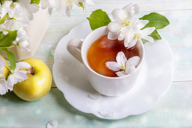 Weiße Tasse Tee auf geformter Untertasse mit Blütenapfelbaumblumen und gelbem Apfel auf Holztisch mit Bokeh-Nahaufnahme.