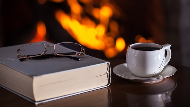 Weiße Tasse Tee oder Kaffee, Gläser und altes Buch nahe Kamin auf Holztisch.