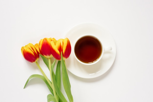Weiße Teetasse mit Untertasse und roten und gelben Tulpen auf weißem Hintergrund Konzept der Liebe und des Frühlings. Flach liegen