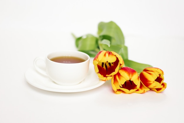 Weiße Teetasse mit Untertasse und roten und gelben Tulpen auf weißem Tisch. Konzept von Liebe und Frühling.