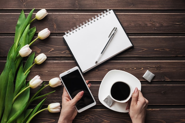 Weiße Tulpen auf einem Holztisch mit einem leeren Notizbuch, einem Smartphone und einem Tasse Kaffee in den Händen der Frauen