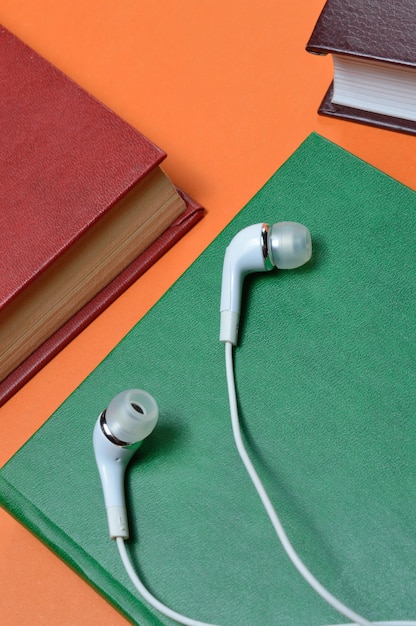 Weiße Vakuumkopfhörer liegen auf Büchern, die auf einem orangefarbenen Hintergrund ausgelegt sind. Hörbuch-Konzept.