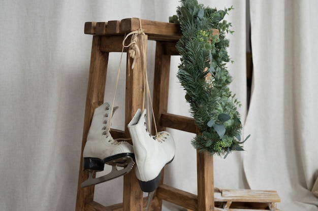 Weiße Vintage Schlittschuhe hängen an Holzhocker, immergrüner Kranz und Stoffhintergrund. Weihnachtsdekoration im Innenbereich.