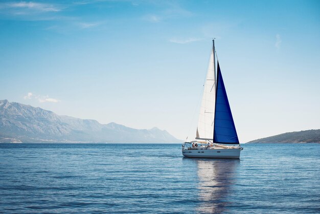 Weiße Yacht mit blauen und weißen Segeln im Meer vor einem Hintergrund aus blauem Himmel und Bergen