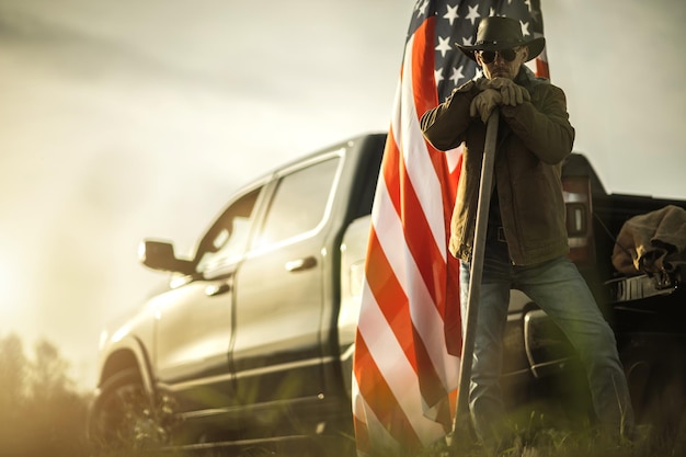 Weißer amerikanischer patriotischer Cowboy-Bauer, der vor seinem Pickup und der amerikanischen Nationalflagge steht