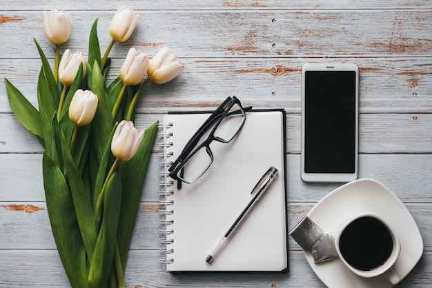 Weißer Blumenstrauß von Tulpen mit Kaffeetasse, Smartphone und leerem Notizbuch