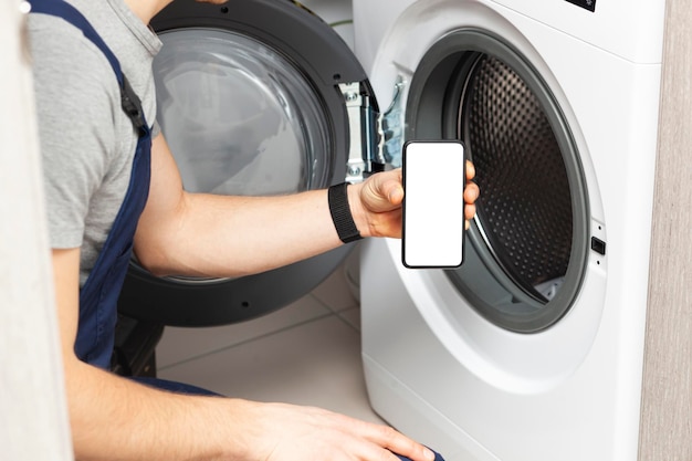 Weißer leerer Bildschirm auf dem Telefon in der Hand des Handwerkers, der gekommen ist, um die Waschmaschine zu reparieren Smartphone-Modell mit leerem Platz Platz für Text auf dem Telefonbildschirm