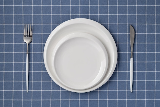Weißer leerer Teller und Besteck auf einer blauen Tischdecke mit kariertem Muster Tischansicht oben Flaches Lay-Design Keramikutensilien zum Servieren von Geschirr Gabel und Messer