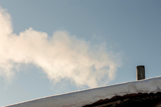 Weißer Rauch aus einem alten Schornstein auf dem mit Schnee bedeckten Dach vor blauem Himmel