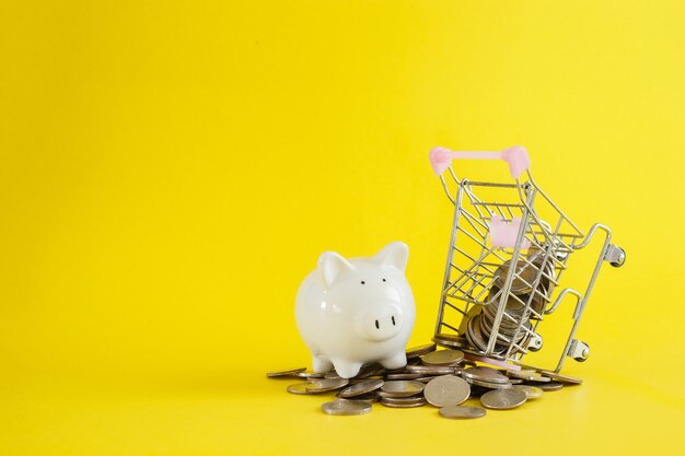 Weißer Schweinchen-Einkaufswagen mit Münzen thailändischer Baht auf gelbem Hintergrund, Spar- oder Investitionskonzept.