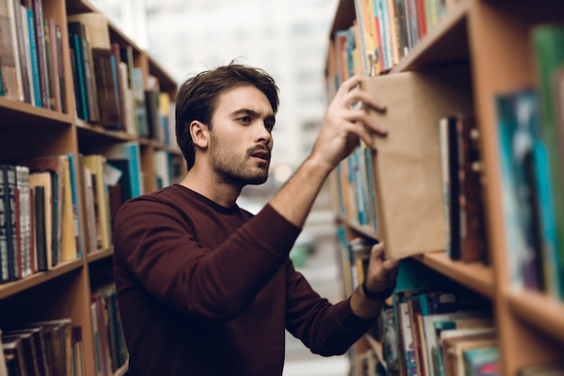 Weißer Student im Pullover mit Büchern im Gang der Bibliothek.