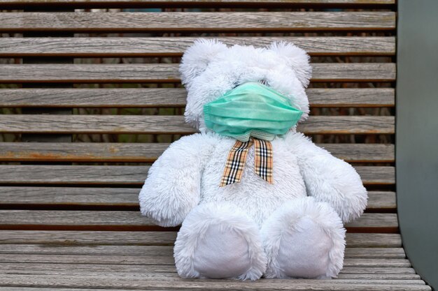 Weißer Teddybär mit schützender medizinischer Gesichtsmaske sitzen allein auf einer hölzernen Parkbank. Krankes Konzept Coronavirus.