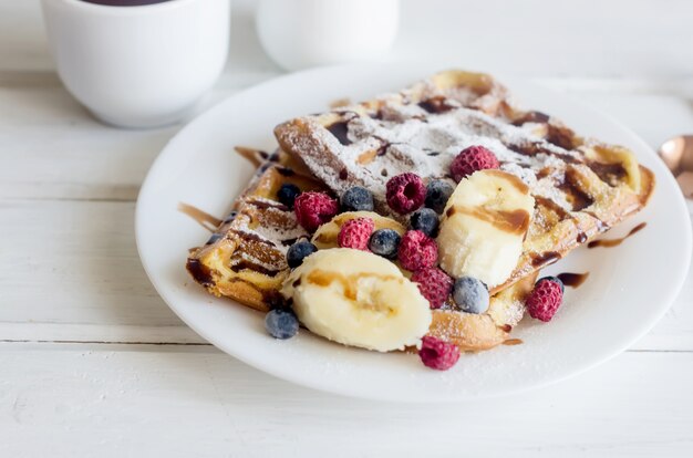 Weißer Teller mit belgischen Waffeln mit Zuckerpulver, gefrorenen Beeren, Bananen-Karamell-Sauce