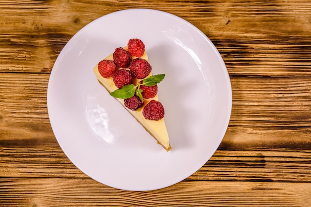 Weißer Teller mit Käsekuchen New York, Minzblatt und Himbeeren auf rustikalem Holztisch. Ansicht von oben