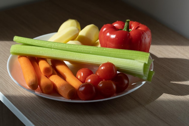 Weißer Teller mit Kirschtomaten, Karotten, Selleriepaprika und Kartoffeln im Sonnenlicht auf Holztisch