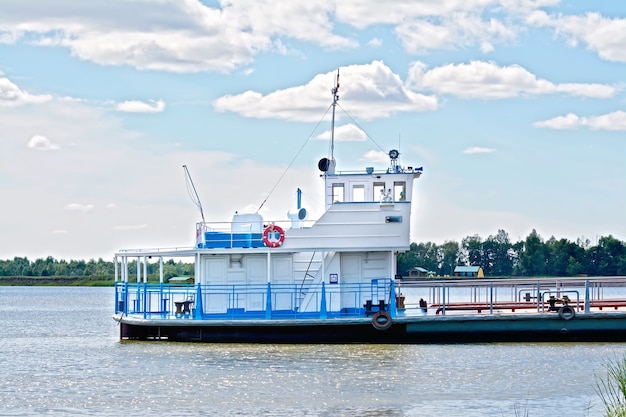 Weißer und blauer Fährenschlepper auf dem Hintergrund von Wasser, Bäumen, blauem Himmel und weißen Wolken