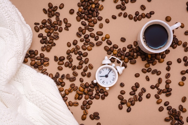 Weißer Wecker, eine Tasse schwarzen Kaffee, ein gemütlicher Pullover und Kaffeebohnen auf beigem Hintergrund. Machen Sie eine Pause von der Arbeit, um sich auszuruhen. Ansicht von oben