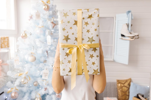 Weißer Weihnachtsbaum verziert mit silbernen goldenen rosa Weihnachtskugeln und schönen Glaskugeln auf funkelnden und feenhaften weißen blauen Zweigen, dekorativen Lichtern, Kopienraum. Foto, Hand hält Geschenkbox