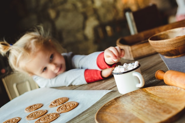 Weißes Baby im traditionellen Schlafanzug nimmt Marshmallows aus einer Tasse Kakao.